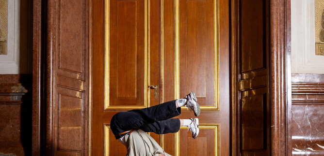 A person dances in a challenging position on a smooth floor. In the background, elegant wooden doors can be seen. | © Raphael Mittendorfer