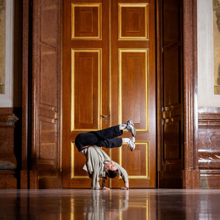 Una persona balla in una posizione impegnativa su un pavimento liscio. Sullo sfondo ci sono porte di legno eleganti. | © Raphael Mittendorfer
