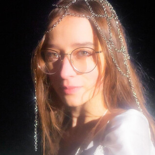 A young woman with glasses and a sparkling headpiece looks directly at the camera. The background is black, which emphasizes her expression. | © Michelle Rassnitzer