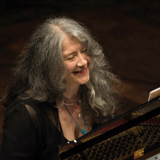 A smiling woman plays the piano. Her long, gray hair falls in curls over her shoulders. | © Adriano Heitman