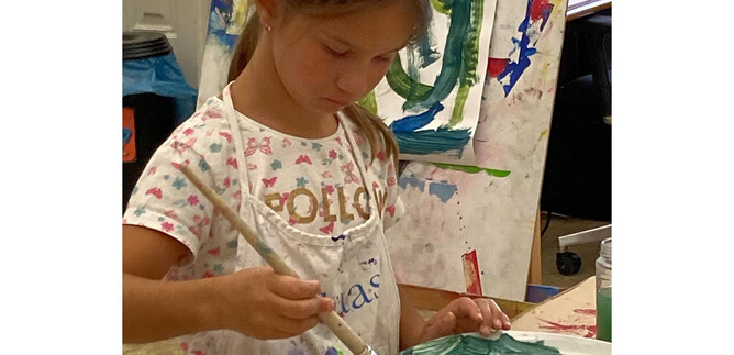 A girl is concentratedly painting with a brush in a bowl. In the background, colorful pictures on canvases can be seen. | © DomQuartier