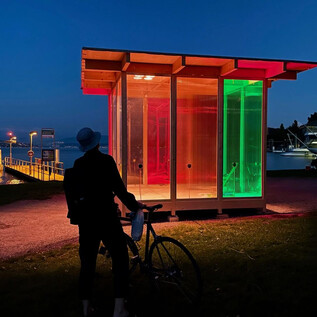 Una moderna cabaña iluminada de vidrio se encuentra a la orilla de un cuerpo de agua. Una persona con una bicicleta observa las luces de colores por la noche. | © DdePerrot
