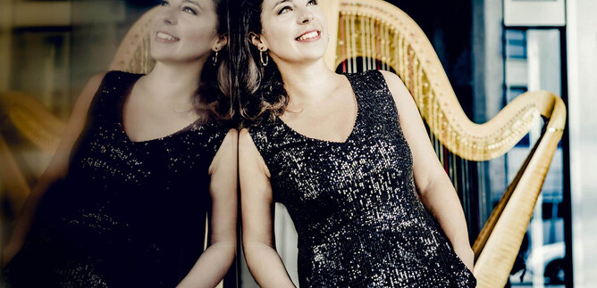 Elegantní žena ve třpytivých šatech se usmívá u zdi. V pozadí je vidět harfa. | © Andrej Grilc