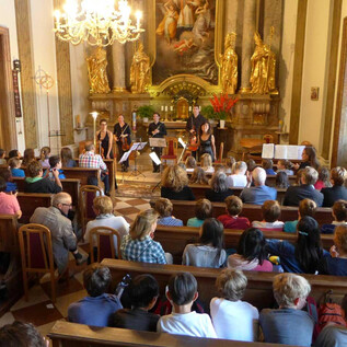 Ein Konzert in einer Kirche mit einer großen Anzahl von Zuhörern. Auf der Bühne spielen mehrere Musiker ihre Instrumente.
