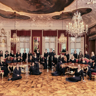 Un gruppo di donne in abiti eleganti è seduto e in piedi in una sontuosa stanza con lampadari. La stanza è riccamente decorata con pareti rosse e un dettagliato affresco sul soffitto. | © Bachchor Salzburg_Foto: Andreas Hechenberger