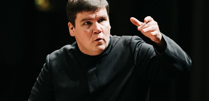 A conductor stands in the center and leads the orchestra. He makes clear gestures with his hand. | © Roberts Blaubuks