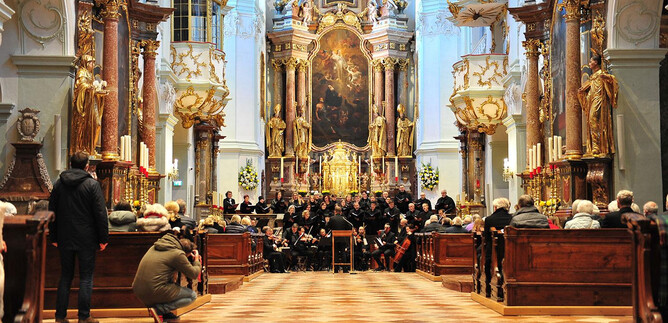 A concert in a magnificent church with a large choir group at the front. The interior design is richly decorated, and many spectators are sitting on the benches. | © P. Alkuin Schachenmayr