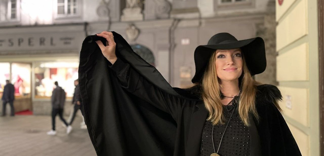 An elegant woman in a black coat and large hat stands in a city street at night. In the background, lights and shop windows can be seen. | © Sabine Rath