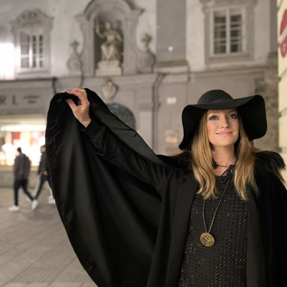 Elegancka kobieta w czarnym płaszczu i dużym kapeluszu stoi na ulicy miasta w nocy. W tle widać światła i witryny sklepowe. | © Sabine Rath