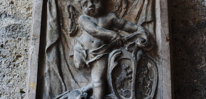 An antique relief of a robe-wearing, winged angel holding a harp. The background features a meticulously designed wall with additional decorative elements. | © Mag. Christoph Koca