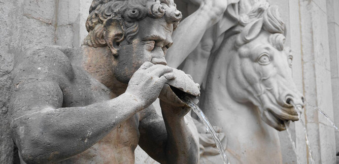 An old statue of a man pouring water from a drinking vessel. In the background, there is a horse with a detailed head. | © Mag. Christoph Koca