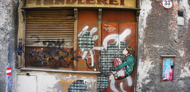 A graffiti-covered shop with the inscription "Töndlerei". A figure sits on the wall, surrounded by colorful artwork. | © Mag. Christoph Koca