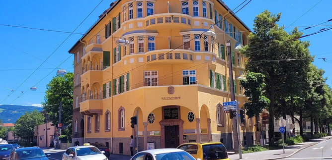 A yellow building with stylish windows and balconies at a street corner. Several cars are passing by, and the surroundings are green and sunny. | © Christoph Koca