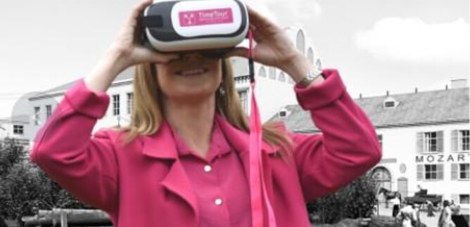 A woman is wearing a virtual reality headset and looking curiously. In the background, historical buildings and people can be seen. | © Sabine Rath