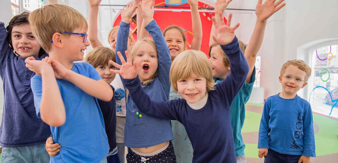 A group of cheerful children stands in a colorful room, raising their hands. They are all wearing blue clothing and radiating joy. | © Eva trifft_Fotografie