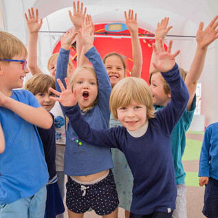 Un groupe d'enfants joyeux se tient dans une pièce colorée et lève les mains. Ils portent tous des vêtements bleus et rayonnent de joie. | © Eva trifft_Fotografie
