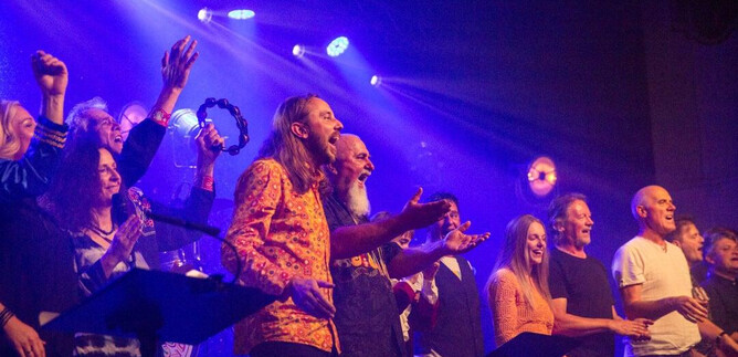 Un grupo de personas canta y celebra en un escenario con luces de colores. La atmósfera es animada y entusiasta. | © Judith Barfuss