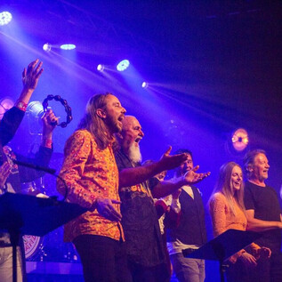 Egy csoport ember énekel és ünnepel egy színpadon színes fényekkel. A hangulat élénk és lelkes. | © Judith Barfuss