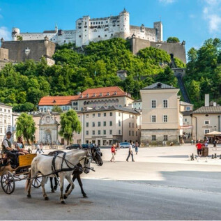 A picturesque scene featuring a horse-drawn carriage and historic architecture. In the background, an impressive fortress and a golden sphere sculpture can be seen. | © TSG