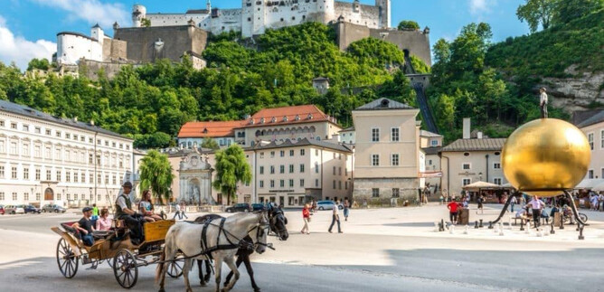 Eine malerische Szene mit einer Pferdekutsche und historischer Architektur. Im Hintergrund ist eine beeindruckende Festung und eine goldene Kugelskulptur zu sehen. | © TSG