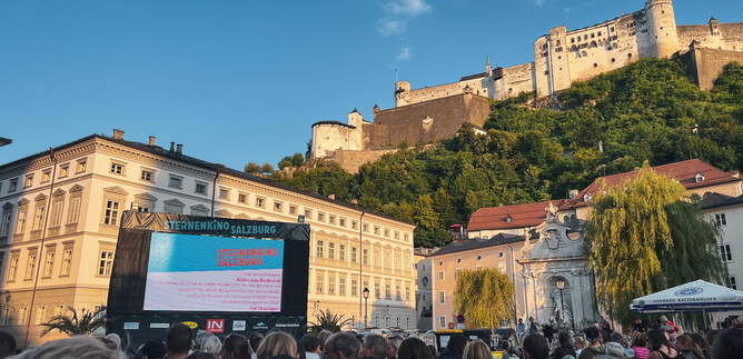 A crowd is sitting in front of a large screen displaying a movie or sports event. In the background, an impressive castle stands on a green hill. | © Tourismus Salzburg GmbH / B. Brunauer