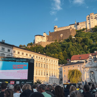 Eine Menschenmenge sitzt vor einer großen Leinwand, die ein Film- oder Sportereignis zeigt. Im Hintergrund steht eine beeindruckende Burg auf einem grünen Hügel. | © Tourismus Salzburg GmbH / B. Brunauer