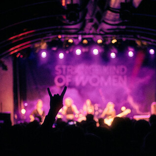 A concert with a band on stage and colorful lights in the background. A fan raises their hand in a sign of excitement. | © Rockhouse Salzburg