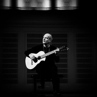 Un musicien est assis sur une chaise et joue de la guitare. La scène est en noir et blanc et dégage une atmosphère calme. | © Laura Pleiffer