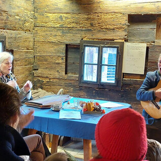 Eine gemütliche Szene in einer Holzhütte. Ein Mann spielt Gitarre, während Frauen begeistert zuhören. | © Salzburger Freilichtmuseum