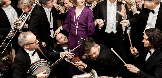 A large group of musicians in elegant evening attire plays various instruments. A woman in a purple dress stands at the center of the scene. | © Erika Mayer