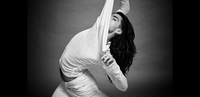 A person in a white top is stretching in a dynamic pose. The background is dark, which puts the focus on the movement and the expression. | © Chris Rogl
