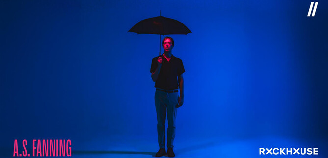 Egy személy áll egy esernyővel egy kék fényben megvilágított háttér előtt. Fekete felsőt visel, és semleges testtartása van. | © Neil Hoare