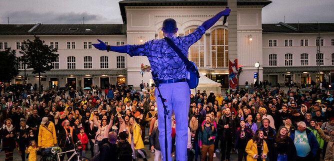 A musician stands on stage and plays in front of a large crowd. The atmosphere is lively and festive. | © Take The A-Train