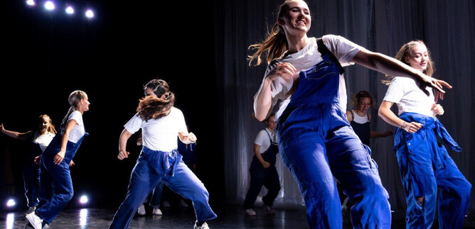 Skupina tanečníků v modrých overalech provádí energickou choreografii na jevišti. Atmosféra je živá s barevnými světly v pozadí. | © Cornelia Pirchner
