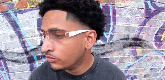 Ein junger Mann mit lockigem Haar und einer modischen Brille steht vor einer bunten Graffiti-Wand. Er blickt nachdenklich zur Seite. | © Timmi Adesanya