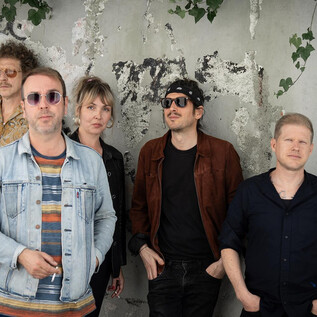 Pięciu muzyków stoi przed szarym murem z graffiti. Uśmiechają się i wyglądają na zrelaksowanych w swoich modnych ubraniach. | © Magdalena Fuchs