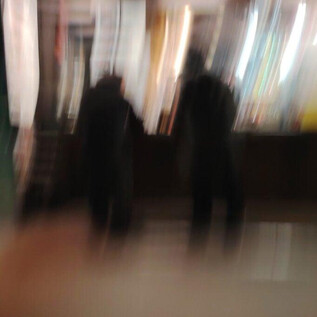 Deux personnes se tiennent floues dans un endroit éclairé. À l'arrière-plan, des fenêtres bien éclairées sont visibles. | © Anna Lindenbaum