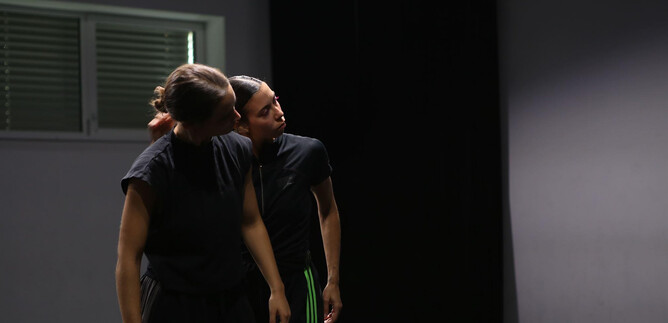 Two dancers in dark clothing stand in a room and seem to have a close interaction. The background is simple and creates a focused atmosphere. | © Dušana Baltic
