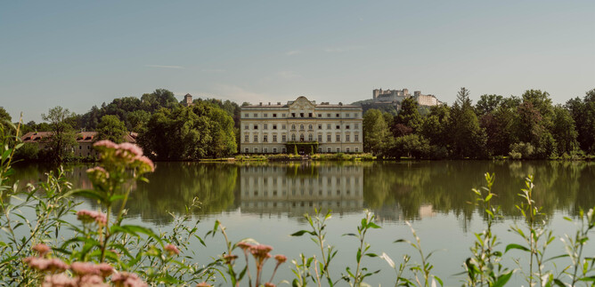 Egy gyönyörű kastély egy nyugodt tó partján, fákkal körülvéve. Előttük színes virágok nyílnak. | © TSG Tourismus Salzburg GmbH, Patrick Langwallner
