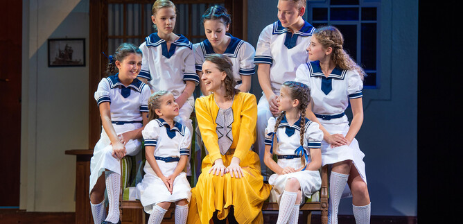 A cheerful scene from a play with children in white uniforms. A woman in a yellow dress sits in the middle and smiles. | © Anna Maria Löffelberger