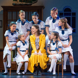 Eine fröhliche Szene aus einem Theaterstück mit Kindern in weißen Uniformen. Eine Frau in einem gelben Kleid sitzt in der Mitte und lächelt. | © Anna Maria Löffelberger