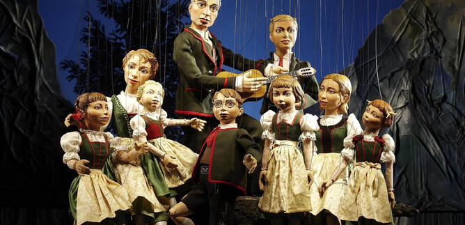 Eine Gruppe von Puppen in traditionellen Trachten steht auf einer Bühne. Eine Puppe spielt Gitarre, während die anderen Puppen um sie herum posieren. | © Salzburger Marionettentheater