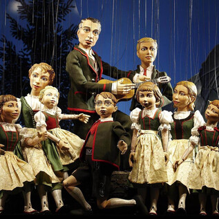 Eine Gruppe von Puppen in traditionellen Trachten steht auf einer Bühne. Eine Puppe spielt Gitarre, während die anderen Puppen um sie herum posieren. | © Salzburger Marionettentheater