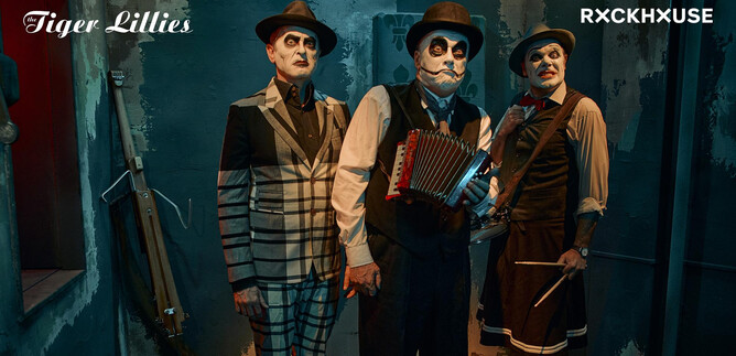 Three men in stylish costumes and with face paint stand in a dimly lit room. One plays an accordion while the others look at the camera. | © Andrey Kezzyn