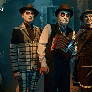 Drei Männer in stilvollen Kostümen und mit Gesichtsmalerei stehen in einem düsteren Raum. Einer spielt ein Akkordeon, während die anderen zur Kamera schauen. | © Andrey Kezzyn