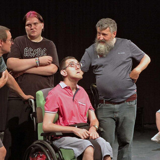 Eine Theaterszene mit mehreren Darstellern, die in einer Diskussion engagiert sind. Einer der Schauspieler sitzt im Rollstuhl, während die anderen ihn umgeben. | © Volxtheaterwerkstatt