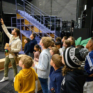 Un gruppo di bambini guarda con interesse a un insegnante che indica un palcoscenico. Sullo sfondo si possono vedere scale e oggetti di scena. | © SLT_Raffael Holzinger