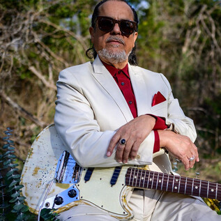 A musician is wearing an elegant white suit and sunglasses. He is sitting outdoors with an electric guitar. | © Lolita Carroll Larriva
