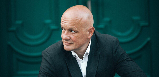 A bald man is wearing a black suit and looking thoughtfully. In the background, green doors are visible. | © Maria Tiefenbacher-Schöndorfer