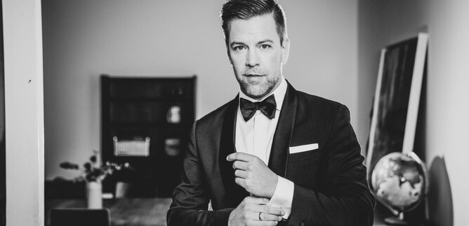 A well-dressed man in a suit with a bow tie stands in a room. The background features furniture and a globe decoration. | © Moritz Künster